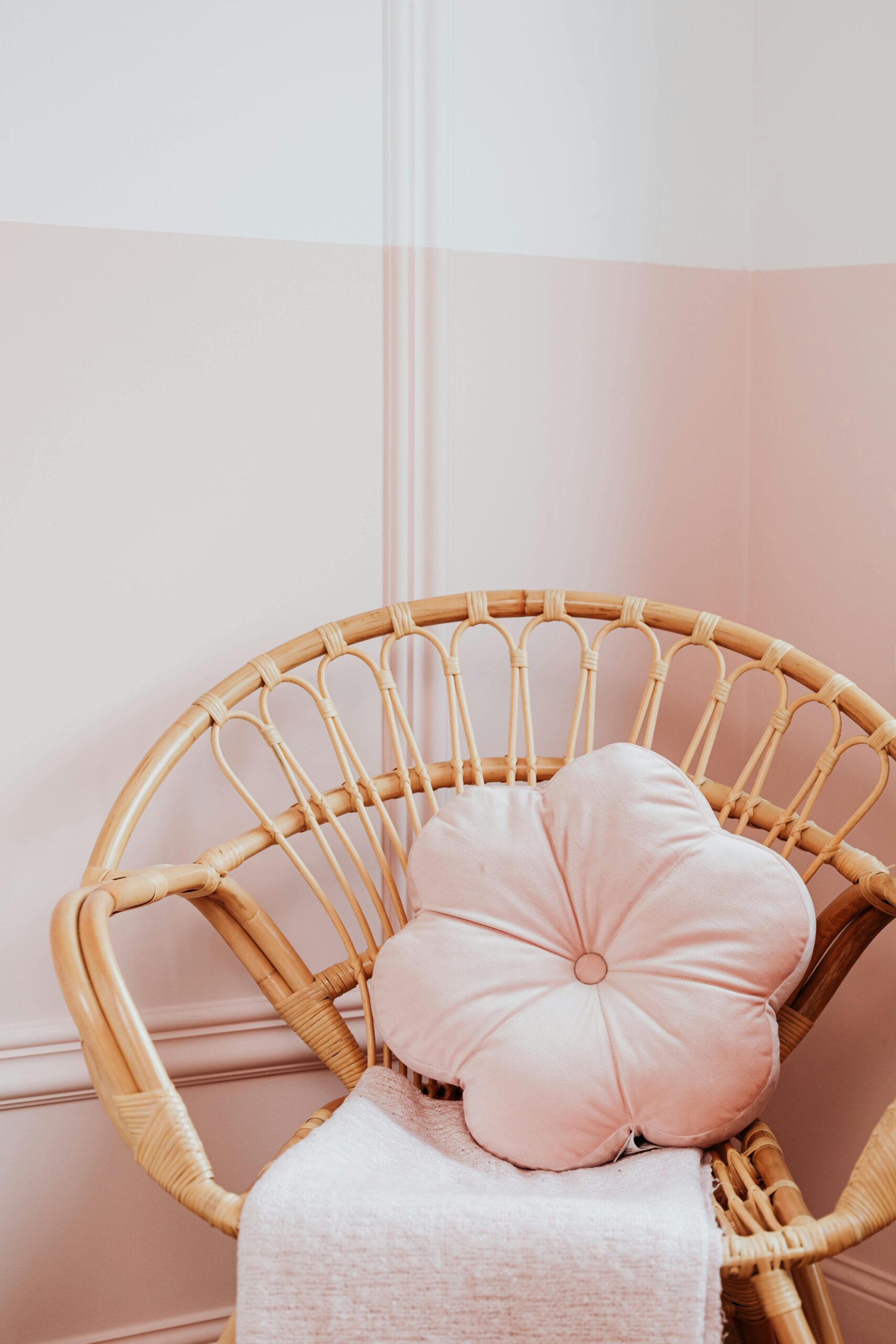 A rattan chair with a pink flower pillow and blanket against a pastel pink wall.